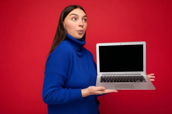 Photo shot of amazed surprised beautiful young brunette woman holding computer laptop looking to the side wearing blue sweater isolated over red wall background