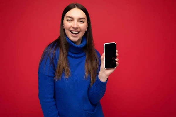 Close-up foto van glimlachende mooie jonge brunette vrouw met casual blauwe trui geïsoleerd over rode achtergrond met lege ruimte in de hand mobiele telefoon en toont smartphone met lege — Stockfoto