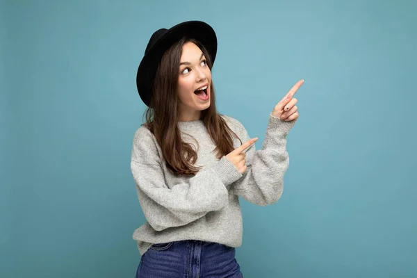 Photo of pretty brunette positive shocked young woman directing fingers to the side with empty space, demonstrating presenting low prices poster, wearing grey sweater and black hat, isolated on blue —  Fotos de Stock