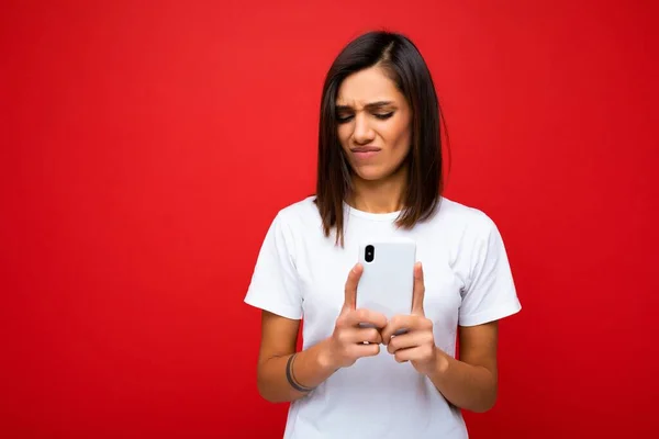 Foto de atractiva insatisfecha molesta joven morena guapa usando casual elegante camiseta blanca posando aislada sobre fondo rojo con espacio vacío sosteniendo en la mano y usando móvil — Foto de Stock