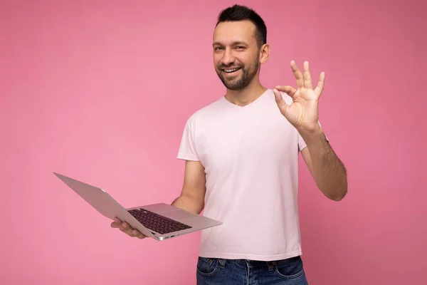 Guapo feliz y divertido morena hombre sosteniendo ordenador portátil y mostrando gesto bien mirando a la cámara en camiseta sobre fondo rosa aislado — Foto de Stock