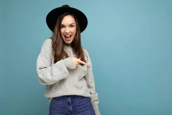 Photo of pretty brunette positive young woman directing finger to the side with empty space, demonstrating presenting low prices poster, wearing grey sweater and black hat, isolated on blue background — Fotografia de Stock