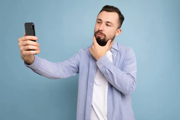 Handsome young man wearing casual stylish clothes standing isolated over background wall holding smartphone taking selfie photo looking at mobile phone screen display — Stock Photo, Image