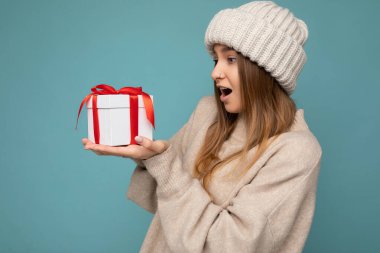 Renkli arka plan duvarından izole edilmiş koyu renk sarışın bir kadının fotoğrafı. Moda kıyafetler giymiş. Elinde hediye kutusu var ve kırmızı kurdeleli şu anki kutuya bakıyor.