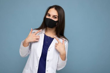 Virüs koruyucu maskeli, yüzü koronavirüse karşı pozitif esmer kadın ve mavi arka planda izole edilmiş beyaz sağlık önlüğü.