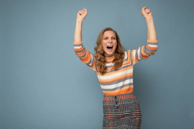 Genç, güzel, mutlu, neşeli, neşeli, neşeli, samimi duyguları olan, şık kıyafetler giyen, mavi arka planda izole edilmiş, boş bir alanda yaşayan ve kazanmayı kutlayan bir kadının fotoğrafı.