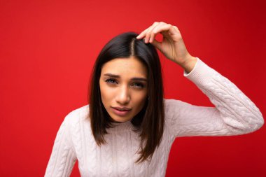 Kısa saç kesimli, samimi duyguları olan, kırmızı arka planda izole edilmiş günlük süveter giyen, fotokopi çeken ve saçlarına dokunan güzel esmer bir kadının yakın plan portresi.