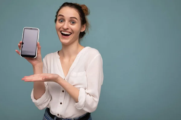 Beautiful positive happy young blonde curly woman wearing casual white shirt and isolated over blue background wall holding phone and showing smartphone with empty dcreen for mockup looking at camera — 스톡 사진