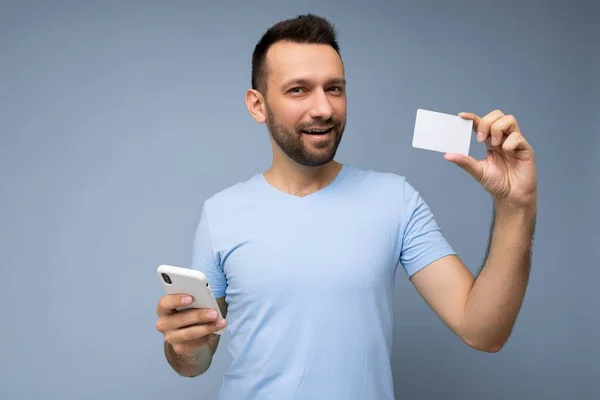 Satisfeito Bonito homem vestindo roupas cotidianas isoladas na parede de fundo segurando e usando telefone e cartão de crédito fazendo pagamento olhando para a câmera — Fotografia de Stock