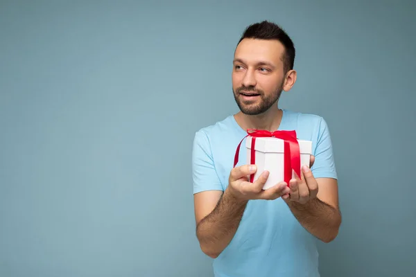 Shot of handsome happy brunette unshaven young man isolated over blue background wall wearing blue t-shirt holding white gift box with red ribbon and looking to the side — 스톡 사진