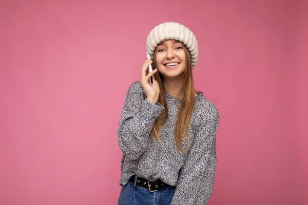 Beautiful positive happy smiling young blonde woman wearing casual grey sweater and beige hat isolated over pink background holding in hand and talking on mobile phone looking at camera — Stock fotografie