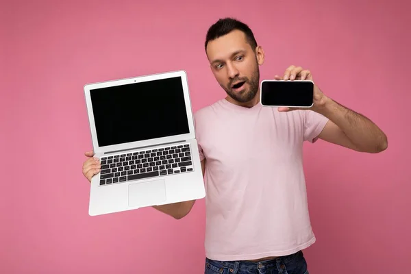 Hombre guapo sosteniendo ordenador portátil y teléfono móvil mirando el teléfono en camiseta sobre fondo rosa aislado —  Fotos de Stock