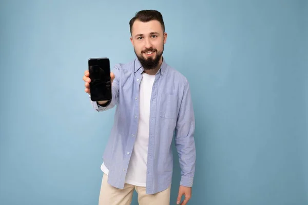 Foto de joven morena guapo serio y guapo sin afeitar hombre con barba que usa camiseta blanca casual y camisa azul posándose aislado sobre fondo azul con espacio vacío sosteniendo en la mano móvil — Foto de Stock