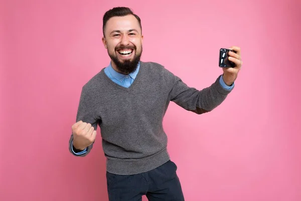 Overjoyed happy handsome young brunette unshaven man with beard wearing everyday grey sweater and blue shirt isolated on background wall holding smartphone watching videos via mobile phone looking at — 스톡 사진