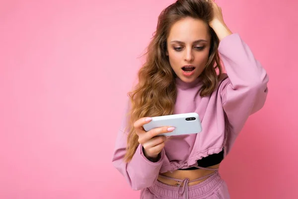 Linda surpreso mulher jovem loira usando capuz rosa isolado sobre fundo rosa com espaço de cópia segurando e usando telefone celular olhando para a exibição do dispositivo — Fotografia de Stock