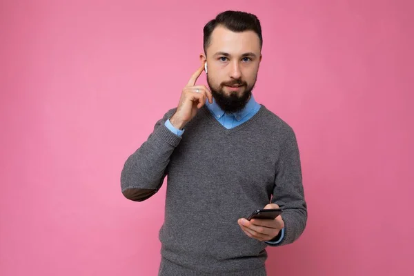 회색 스웨터를 입고 파란 셔츠를 입은 잘생긴 갈색 턱수염의 젊은 남자가 손에 빈 공간을 잡고 휴대폰을 사용하여 온라인으로 대화하는 모습은 분홍색 배경에 고립되어 있다. — 스톡 사진