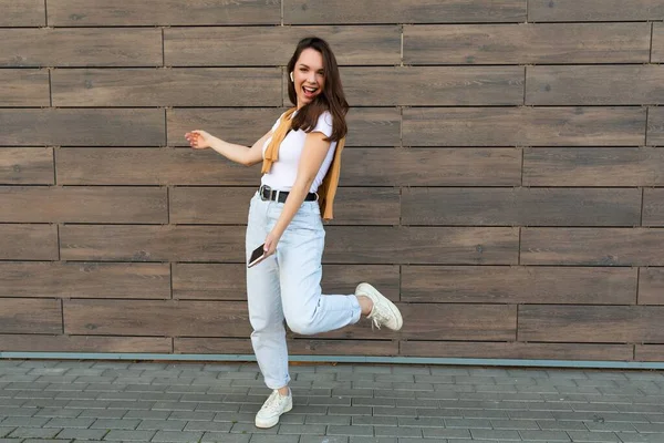 Full length body size photo of pretty brunet young happy woman wearing casual clothes and listening to music via wireless earphones standing in the street holding and using mobile phone looking at — Stock Photo, Image