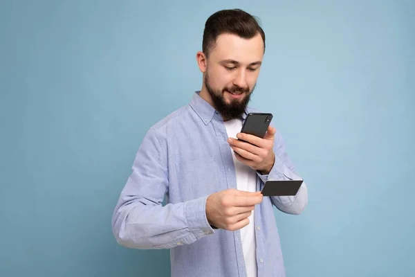 Jóképű férfi visel mindennapi ruhát elszigetelt háttér fali gazdaság és a telefon és a hitelkártya fizetés nézi okostelefon képernyő — Stock Fotó