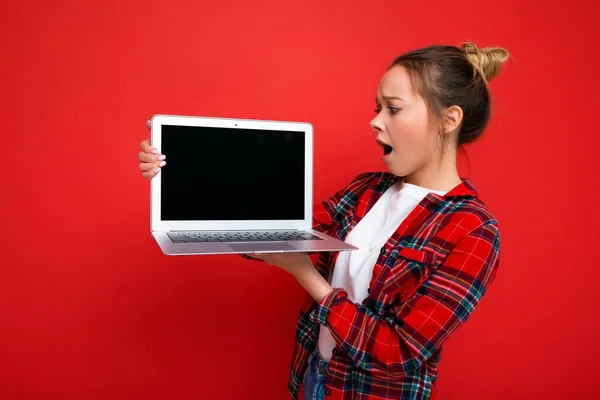 Foto de bela chocado mulher jovem surpreendente segurando laptop vestindo camisa vermelha olhando monitor de exibição isolado no fundo vermelho — Fotografia de Stock
