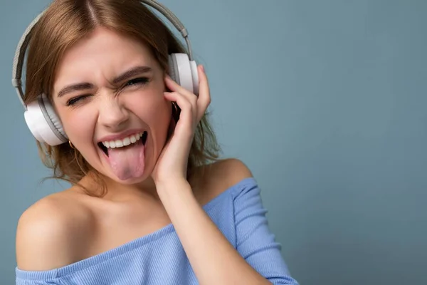 Closeup portrait of attractive emotional young blonde woman wearing blue crop top isolated on blue background wearing white wireless bluetooth headphones listening to cool music and having fun looking — Stock Photo, Image