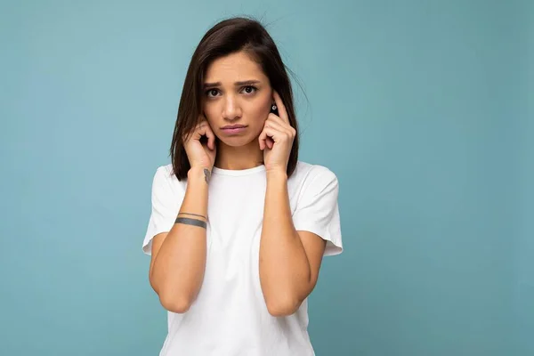 Retrato de triste triste joven morena bastante agradable con emociones sinceras que lleva una camiseta blanca casual para maqueta aislada sobre fondo azul con espacio para copiar y cubrir las orejas con los dedos — Foto de Stock