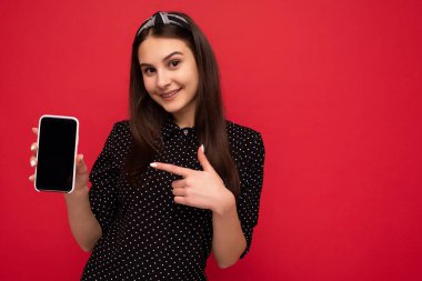 Güzel, gülümseyen, şık giyinen, arka planda izole bir şekilde duran akıllı telefonlu ve elinde boş ekran görüntüsü olan güzel bir gencin fotoğrafı.