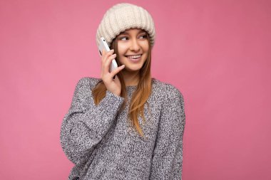 Çekici, pozitif, mutlu, gri süveter ve bej şapka giyen genç bir kadın. Pembe arka planda izole edilmiş. El ele tutuşup cep telefonuyla konuşuyor.