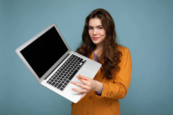 Foto de Hermosa mujer joven feliz seria sosteniendo computadora portátil mirando a la cámara con chaqueta amarilla aislada sobre fondo de pared azul —  Fotos de Stock