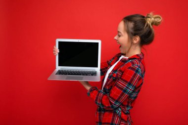 Yan profil fotoğrafı kırmızı arka planda izole edilmiş monitöre bakan kırmızı tişörtlü güzel, şaşkın genç bir kadın.