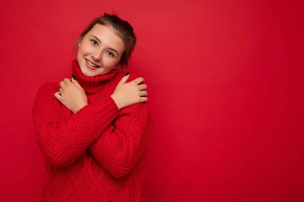Ελκυστικό χαριτωμένο χαμογελαστό νεαρή γυναίκα φορώντας ζεστό κόκκινο πουλόβερ απομονώνονται πάνω από το κόκκινο φόντο τοίχο κρατώντας τα χέρια στο στήθος κοιτάζοντας κάμερα — Φωτογραφία Αρχείου