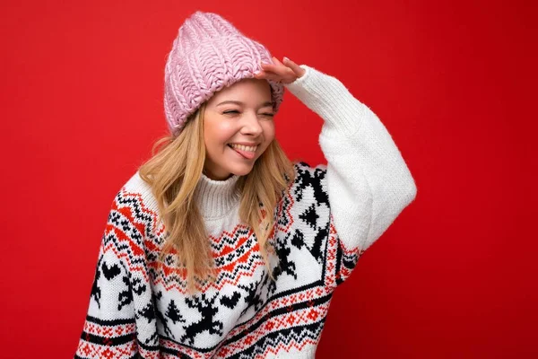 Foto de joven positiva feliz alegre atractiva mujer rubia con emociones sinceras usando sombrero de punto y jersey de invierno cálido aislado sobre fondo rojo con espacio vacío y mirando a un lado con —  Fotos de Stock
