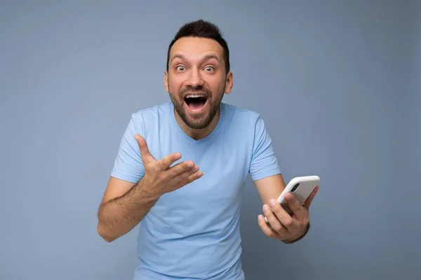 Потрясенный красивый молодой бородатый мужчина в повседневной синей футболке на синем фоне, держащий и использующий мобильную связь онлайн в Интернете, глядя на gadjet дисплей — стоковое фото