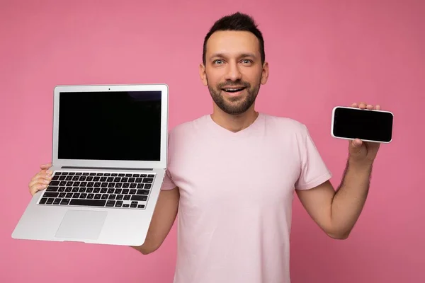 Bonito morena homem segurando computador portátil e telefone celular olhando para a câmera em t-shirt no fundo rosa isolado — Fotografia de Stock