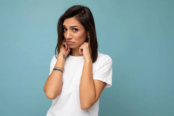 Portret van verdrietig beledigd verdrietig jong vrij mooi brunette vrouw met oprechte emoties dragen casual wit t-shirt voor mockup geïsoleerd op blauwe achtergrond met kopieerruimte en het bedekken van oren met — Stockfoto