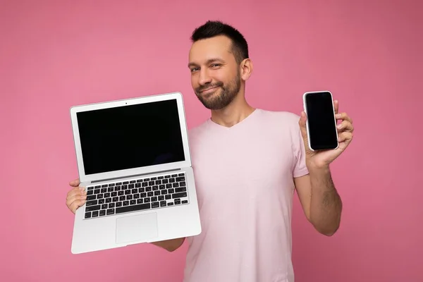 Hombre guapo morena sosteniendo ordenador portátil y teléfono móvil mirando a la cámara en camiseta sobre fondo rosa aislado — Foto de Stock
