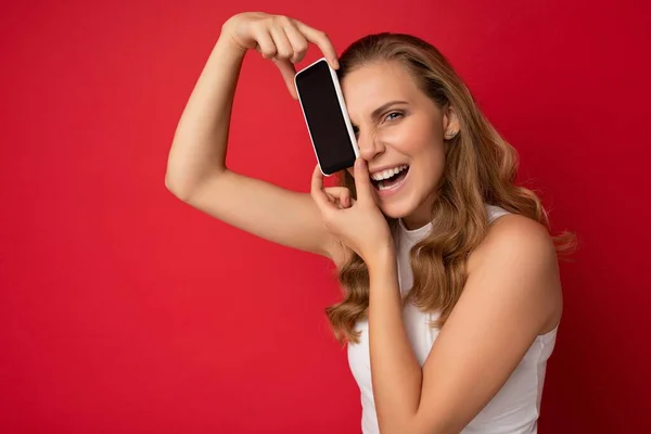 Shot of pretty emotional funny young blonde female person wearing white t-shirt isolated on red background with copy space holding smartphone showing phone in hand with empty screen for cutout looking — Stock Photo, Image