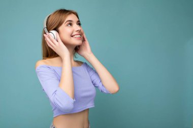 Mavi ekin bluz giyen güzel, pozitif gülümseyen sarışın bir kadının mavi arka plan duvarına izole edilmiş beyaz kablosuz bluetooth kulaklık takarak havalı müzik dinleyen bir kadının yan profil fotoğrafı.