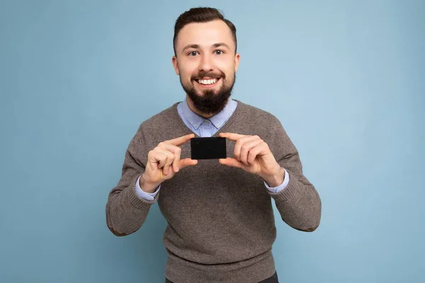 Bonito sorridente morena barbuda homem vestindo camisola cinza e camisa azul isolado na parede de fundo segurando cartão de crédito olhando para a câmera — Fotografia de Stock