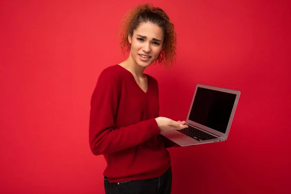 Portret de aproape de frumos uimit surprins uimit întuneric blonda femeie care deține laptop calculator uitându-se la tastarea camerei pe tastatură purtând pulover roșu izolat pe fundal de perete roșu — Fotografie, imagine de stoc