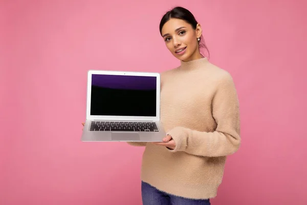 Gyönyörű mosolygós fiatal nő sötét hajjal nézi kamera gazdaság számítógép laptop üres monitor képernyő, mock up és másolja tér visel világos színű pulóver elszigetelt rózsaszín falon — Stock Fotó