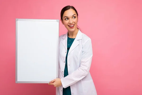 Porträt einer attraktiven hübschen jungen positiven brünetten Frau in medizinischem weißen Mantel mit weißer Magnettafel für Attrappe isoliert über rosa Hintergrund mit Kopierraum — Stockfoto