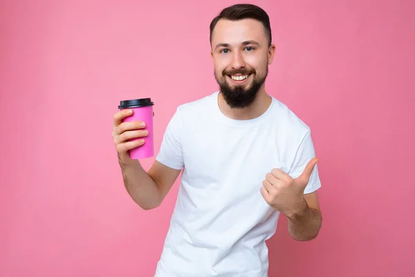 수염을 기르고 흰색 티셔츠를 입고 있는 잘생긴 미소를 짓고 있는 젊은 남성의 사진이 분홍색 배낭 벽에 고립되어 있고 종이 커피 잔을 들고 있으며 카메라를 보고 있다. — 스톡 사진