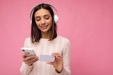 Güzel, gülümseyen, esmer, pembe süveter giyen bir kadının fotoğrafı. Pembe arka plan duvarında izole edilmiş, beyaz bluetooth kulaklık takıyor, müzik dinliyor ve cep telefonu kullanıyor.