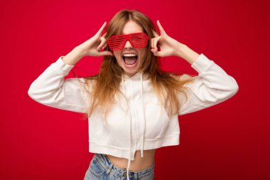 Güzel, pozitif sarışın bir kadının, günlük kıyafetler giydiği ve renkli arka plan duvarından izole edilmiş şık gözlüklü bir şekilde kameraya bakıp eğlendiği bir fotoğraf.