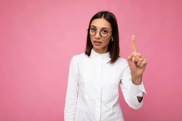 Fotografering av vackra sexig strikt ung brunett kvinna bär casual kläder och snygga optiska glasögon isolerade över färgglada bakgrund tittar på kameran och visar ett finger — Stockfoto