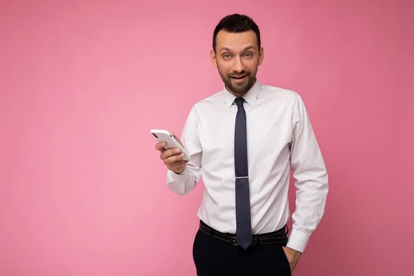 잘 생긴 잘생긴 남자가 흰색 셔츠를 입고 핑크 색 배경에서 고립된 사진 손에 빈 공간을 잡고 카메라를 보는 휴대 전화 메시지를 사용하는 모습 — 스톡 사진