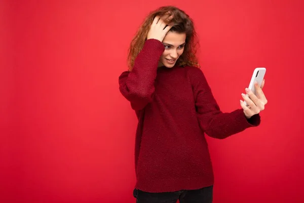 Charmante jonge vrouw met krullend haar dragen donker rode trui geïsoleerd op rode achtergrond muur houden en met behulp van smart phone kijken naar telefoonscherm en het hebben van videogesprek — Stockfoto