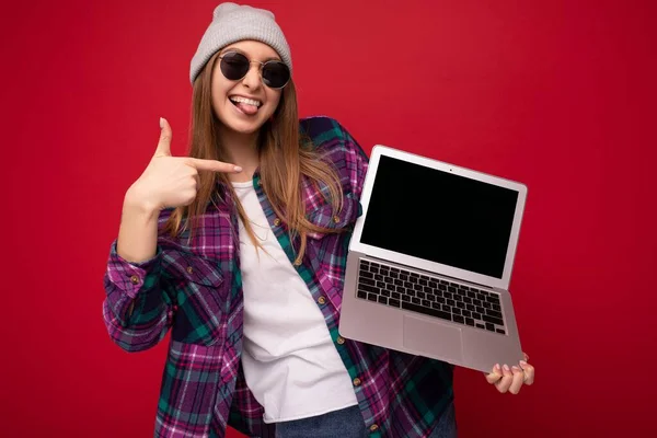 Împușcat de râs amuzant zâmbind tânără femeie care deține laptop-ul computerului purtând pălărie de ochelari de soare și cămașă colorată uitându-se la camera care arată limba și arătând spre ecranul netbook izolat peste peretele roșu — Fotografie, imagine de stoc