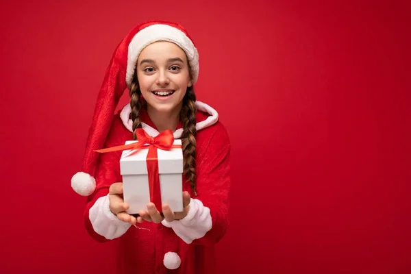 Foto av vacker glad leende brunett flicka isolerad över röd bakgrund vägg bär Santa claus kostym håller vit presentförpackning med rött band och tittar på kameran — Stockfoto