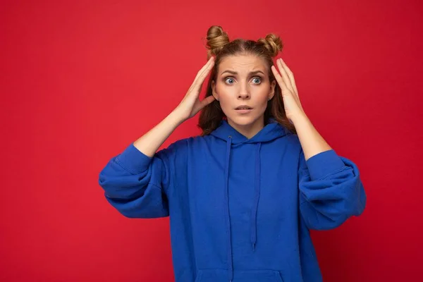 Foto de emocional surpreendido surpreendido jovem bela mulher loira com dois chifres com emoções sinceras vestindo hipster capuz azul brilhante isolado sobre fundo vermelho com espaço livre — Fotografia de Stock